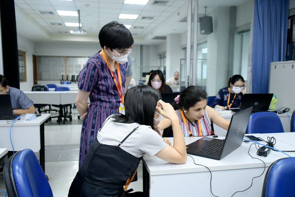 A colour photo of Polly Yap seated at a computer, with a peer participant (also seated) studying the computer screen. A course instructor is standing my their workstation, engaging in direct discussion with Polly and her colleague. Photo credit: Mahidol University